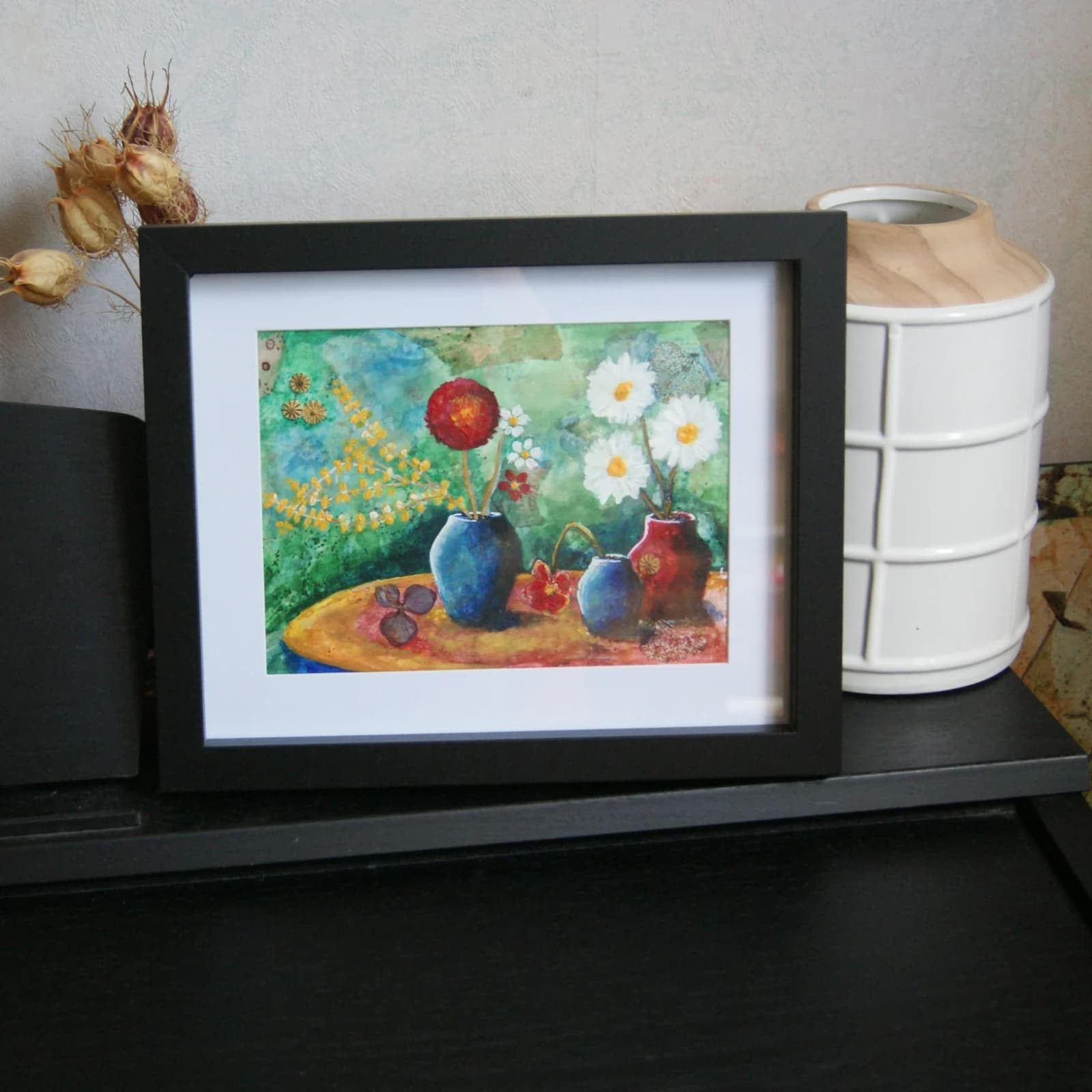 Peinture florale avec vases colorés encadrée en noir sur un meuble à côté d'un vase blanc.
