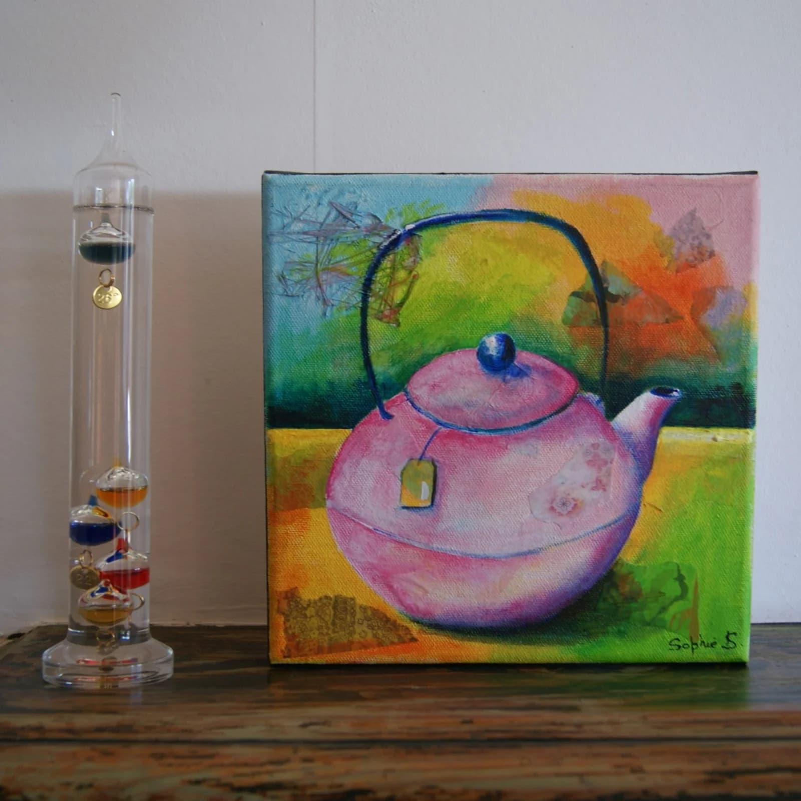 Tableau sur toile d'une théière rose posé sur un meuble en bois à côté d'un thermomètre en verre