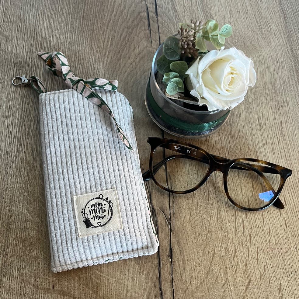 Étui à lunettes en velours côtelé beige clair posé à plat à côté d'une monture et d'une rose blanche.