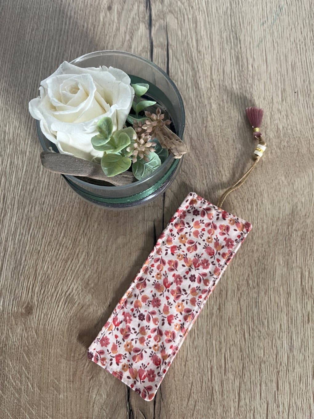 Marque-page rectangulaire en tissu fleuri rose avec cordonnet et pompon posé sur une table en bois