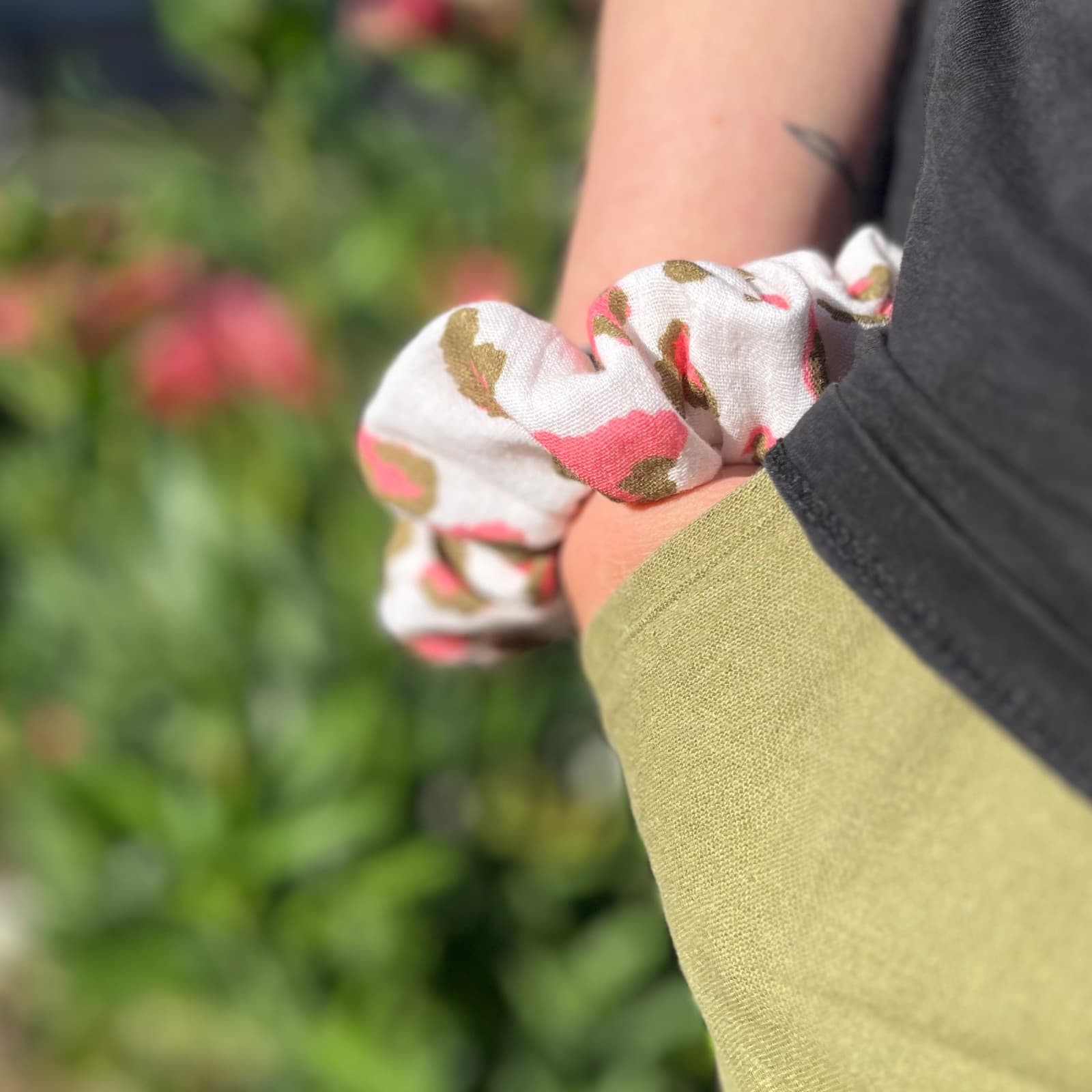 Chouchou froncé blanc à motifs léopard rose et kaki porté au poignet, sur fond de verdure.