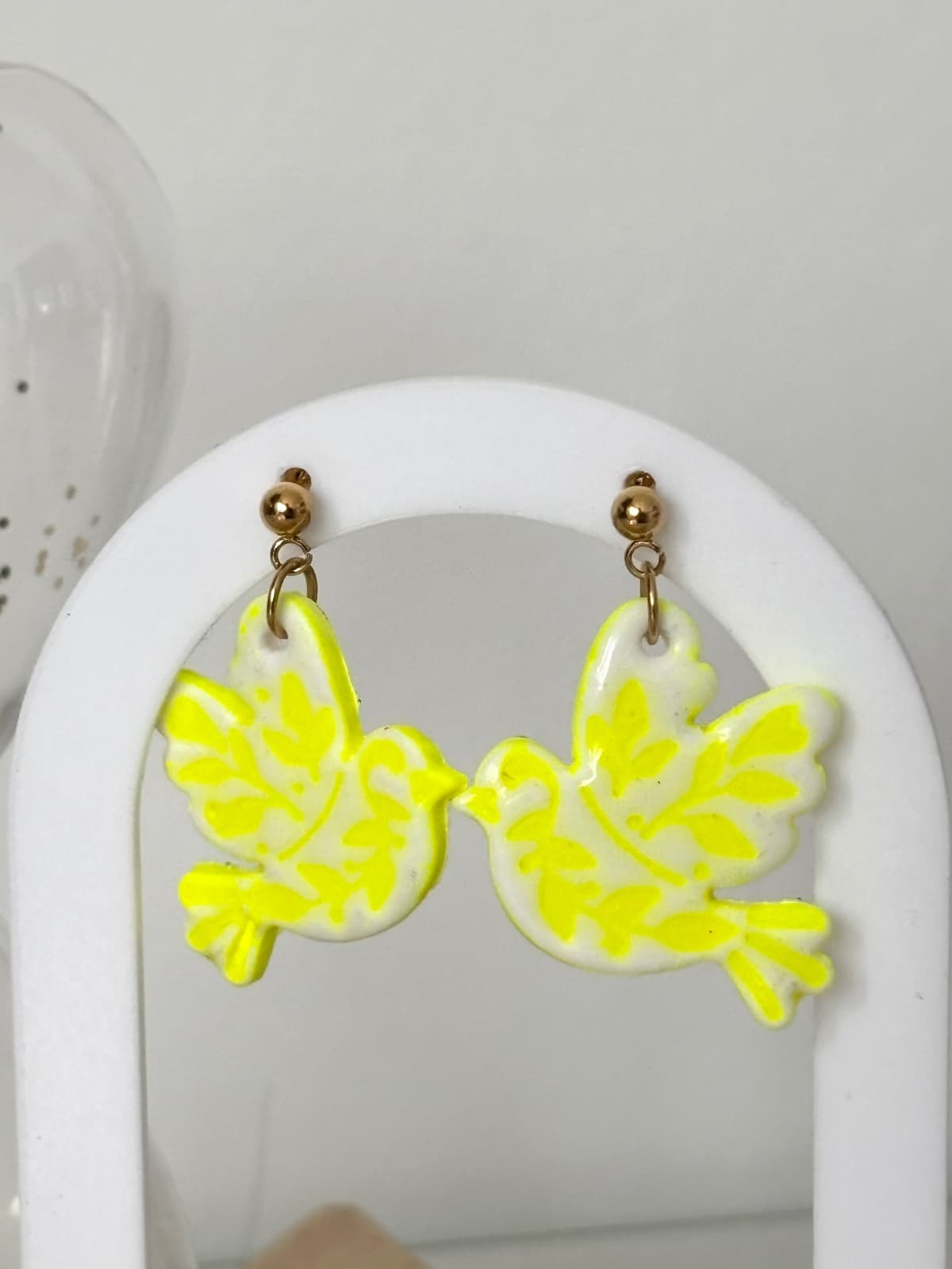 Paire de boucles d’oreilles oiseau blanches et jaune néon suspendues à un présentoir blanc.