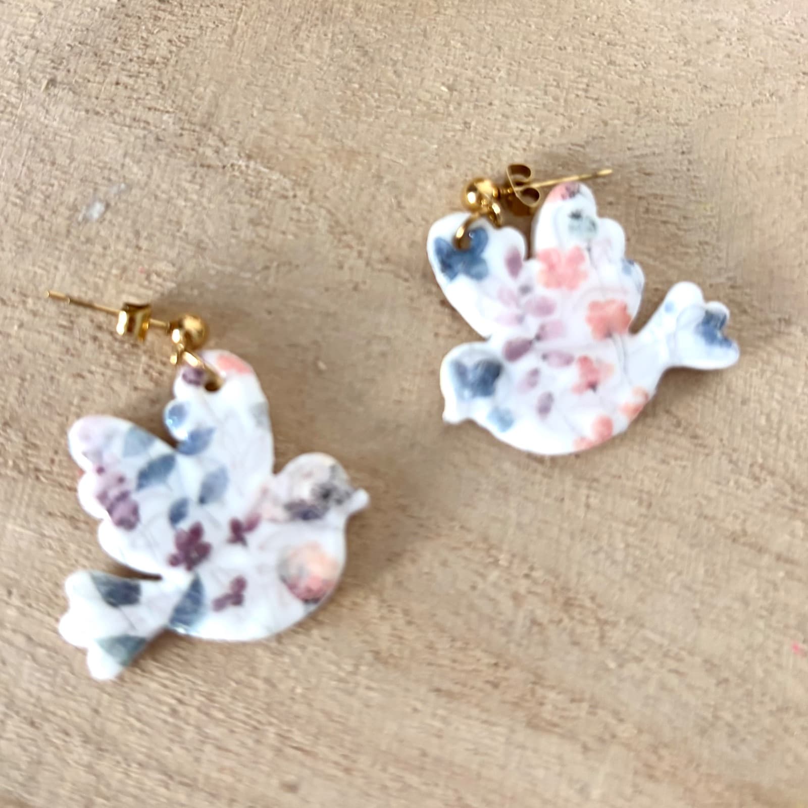 Paire de boucles d'oreilles oiseau fleuries aux tons roses et bleus posée sur une surface en bois.