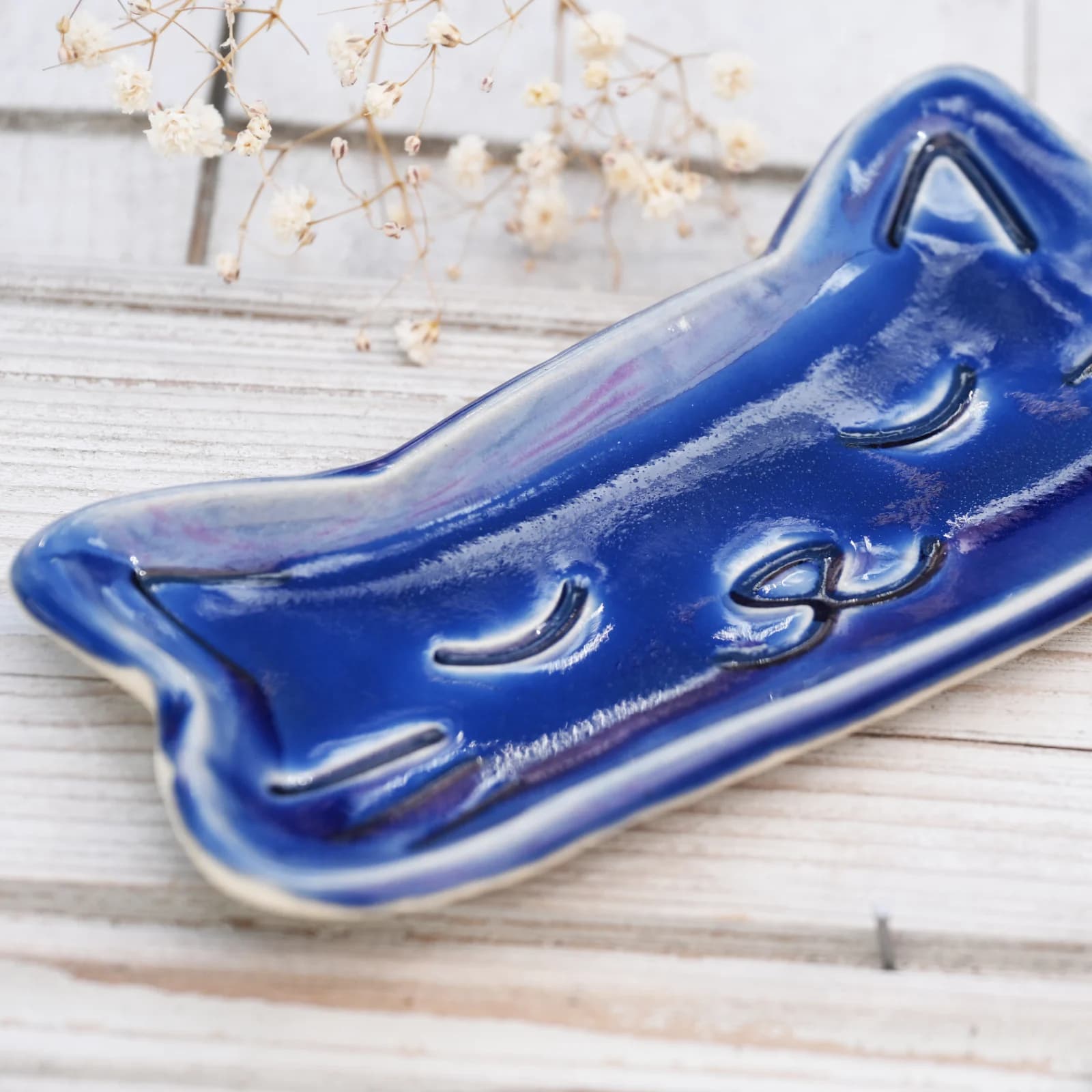 Plateau en céramique bleu brillant en forme de tête de chat aux yeux clos, posé sur une surface en bois clair.