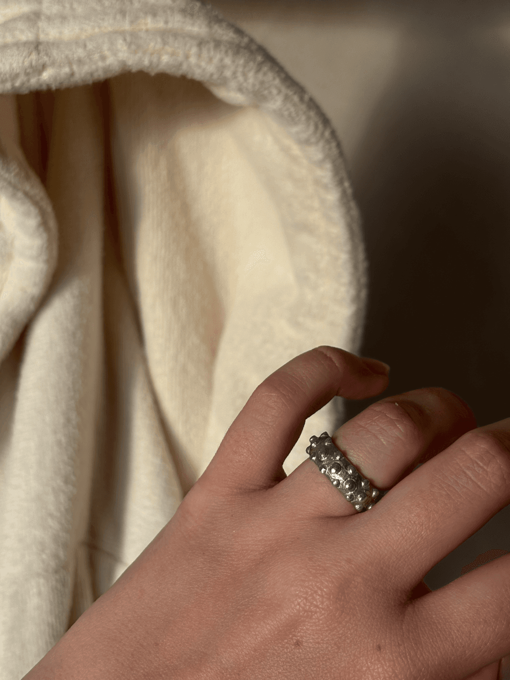 Bague en étain texturée de petites sphères en relief portée à la main devant un tissu écru.